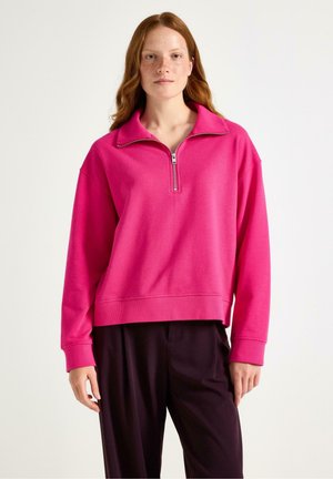 Jongeman met rood haar en sproeten, gekleed in een felle roze half-rits pullover en donkere losse broeken, tegen een eenvoudige lichte achtergrond.