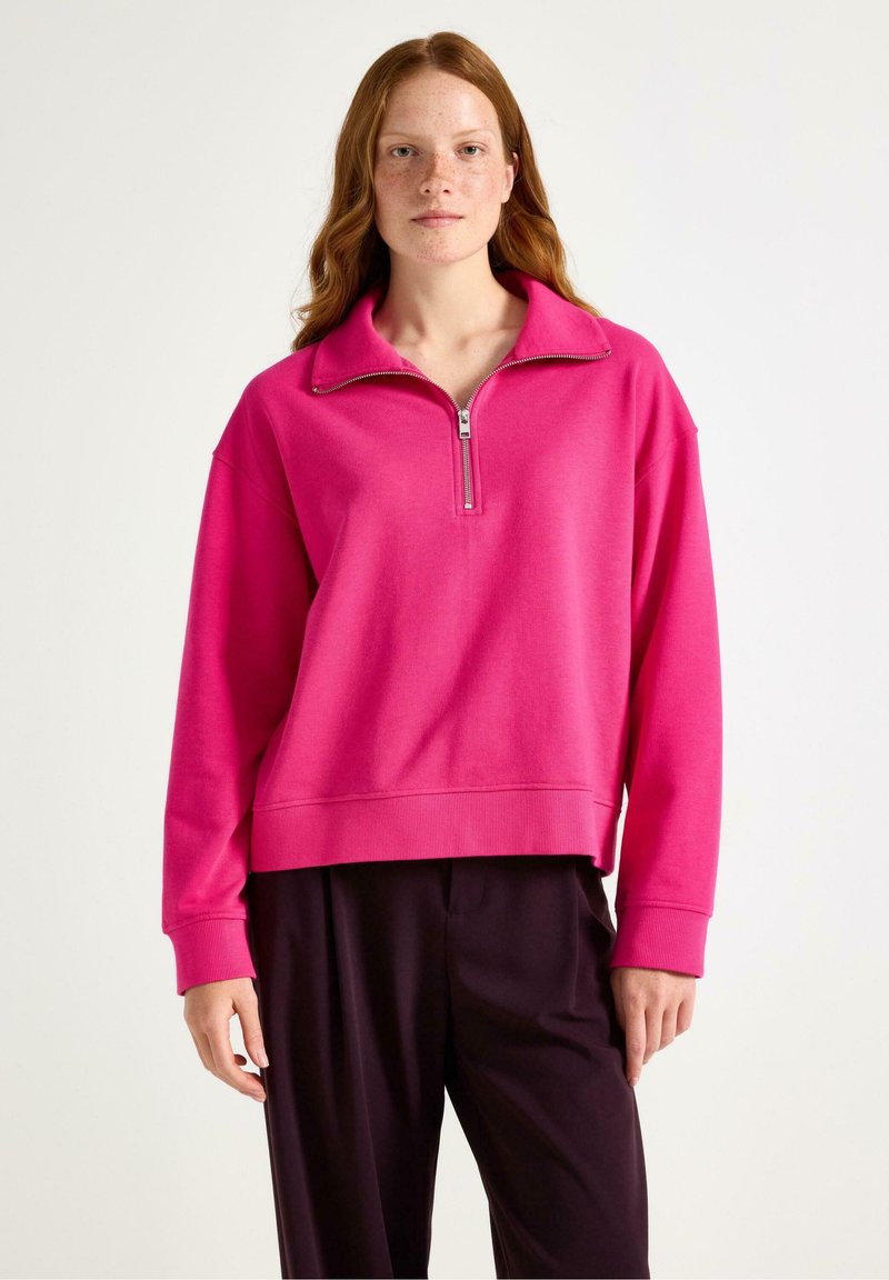 Ung kvinde med rødt hår og fregner iført en lys pink halv-zip trøje og mørke løse bukser mod en ensfarvet lys baggrund.