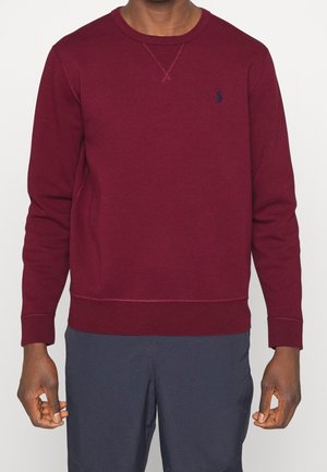 Homme portant un sweat-shirt à col rond à manches longues de couleur bordeaux avec un petit logo bleu marine sur la poitrine gauche et un pantalon bleu marine.