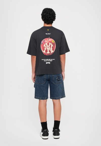 Camiseta negra de manga corta con un gran gráfico de una manzana en la espalda, acompañado de texto y pegatinas coloridas. Combinada con pantalones cortos de mezclilla oscuros.