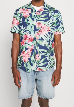 Homme portant une chemise bleu marine à manches courtes avec des motifs floraux roses et feuilles vertes, associée à un short en jean bleu clair.