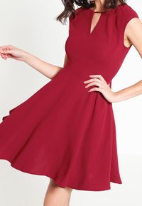 Burgundy ärmlös klänning med nyckelhålskörning, figurnära liv och utsvängd kjol, tillverkad av lätt material med slät textur.