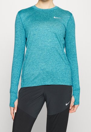 Person trägt ein sportliches langärmeliges Oberteil in Türkis und schwarze Nike-Leggings, steht mit entspannten Händen an den Seiten.