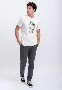 T-shirt en coton blanc avec une illustration de main grise, associé à un jogging gris foncé avec des accents verts et des basket noires.