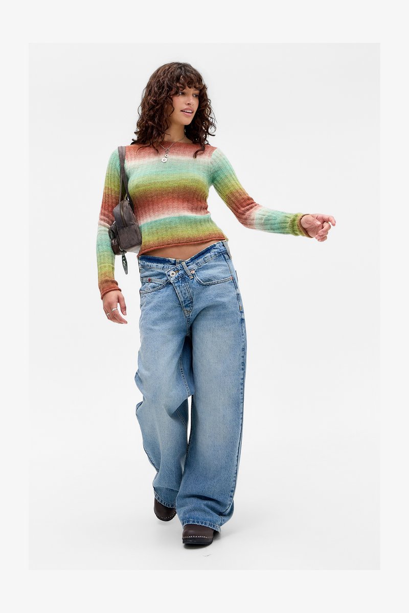 Frau, die selbstbewusst geht, trägt einen mehrfarbigen gestreiften Pullover, weiter geschnittene Jeans mit gekreuztem Bund, braune Schuhe und trägt eine braune Umhängetasche.