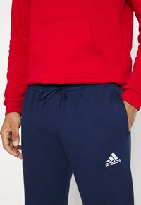 Roter Kapuzenpulli mit einer Fronttasche, kombiniert mit navyblauen Jogginghosen mit elastischem Bund und weißem Adidas-Logo.