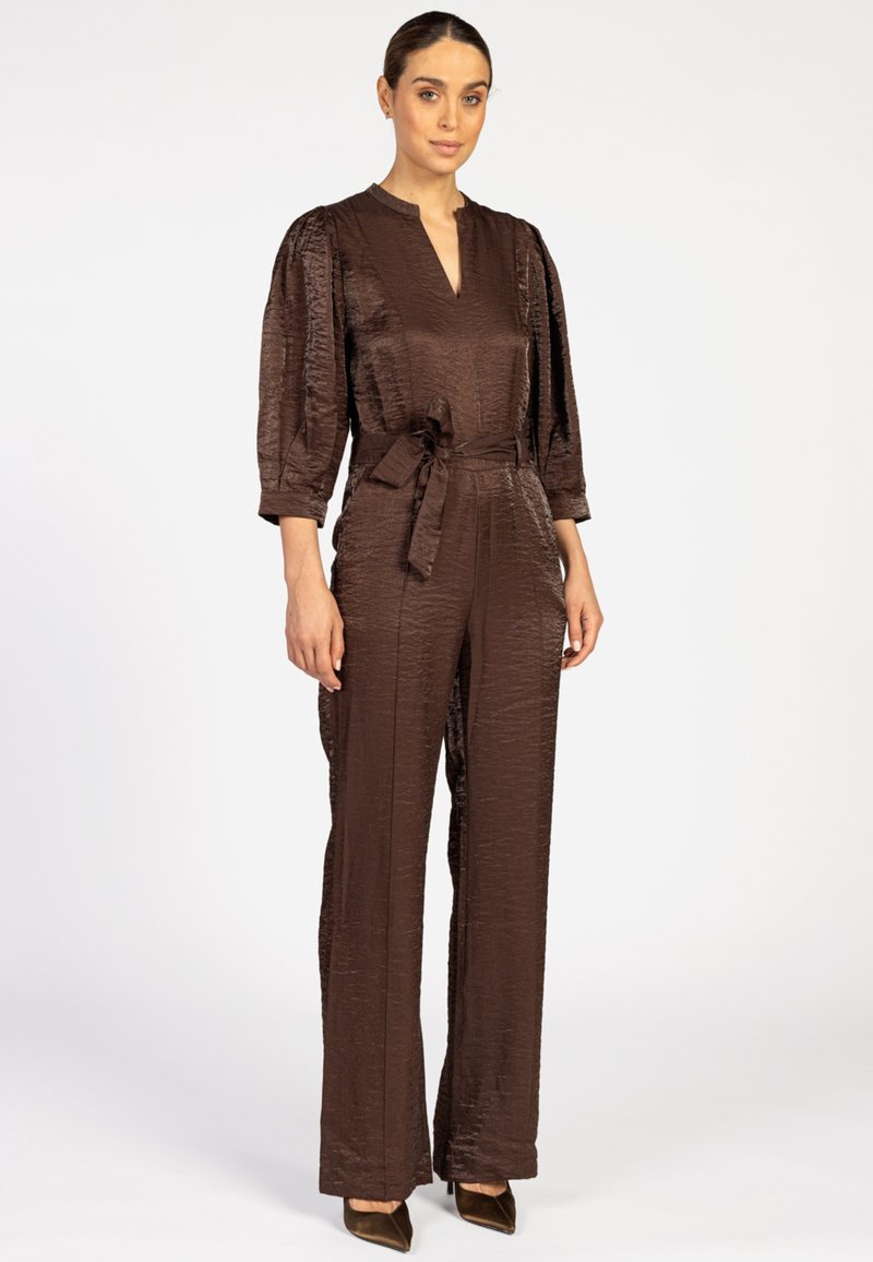 Bruine satijnen jumpsuit met driekwart pofmouwen, een V-hals en een tailleband. Het ontwerp heeft een gestructureerde afwerking en een wijd uitlopende pijp.