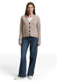 Woman wearing a beige buttoned cardigan over a white top, blue wide-leg jeans, and white sneakers, standing against a white background.