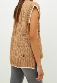 Strik beige sweater med et struktureret mønster, korte ærmer og en hvid bølget kant. Bruges over grå jeans og har en løs pasform.