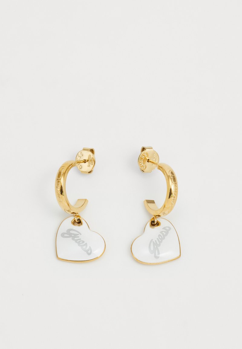 Orecchini a cerchio in oro con un ciondolo a forma di cuore in smalto bianco che presenta la parola "Guess" in grigio. Include un marchio inciso sul cerchio.