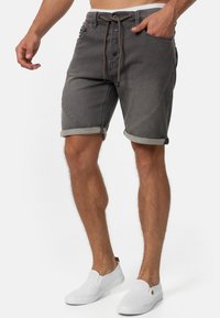 Graue Denim-Shorts mit umgeschlagenen Säumen, vorderer Knopfverschluss und dekorativen seitlichen Akzenten. Getragen mit weißen Slipschuhen.