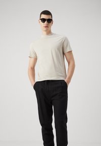 Camiseta beige de manga corta y pantalones negros con cordón. Modelo con gafas de sol negras, de pie con las manos en los bolsillos frente a un fondo neutro.