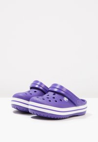 Zoccoli in plastica viola con fori di ventilazione, striature bianche attorno alla suola e una tracolla per una vestibilità sicura. Texture liscia.
