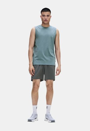 Jeune homme debout portant un débardeur turquoise, un short gris foncé, des chaussettes blanches montantes et des chaussures de sport blanches sur un fond uni.