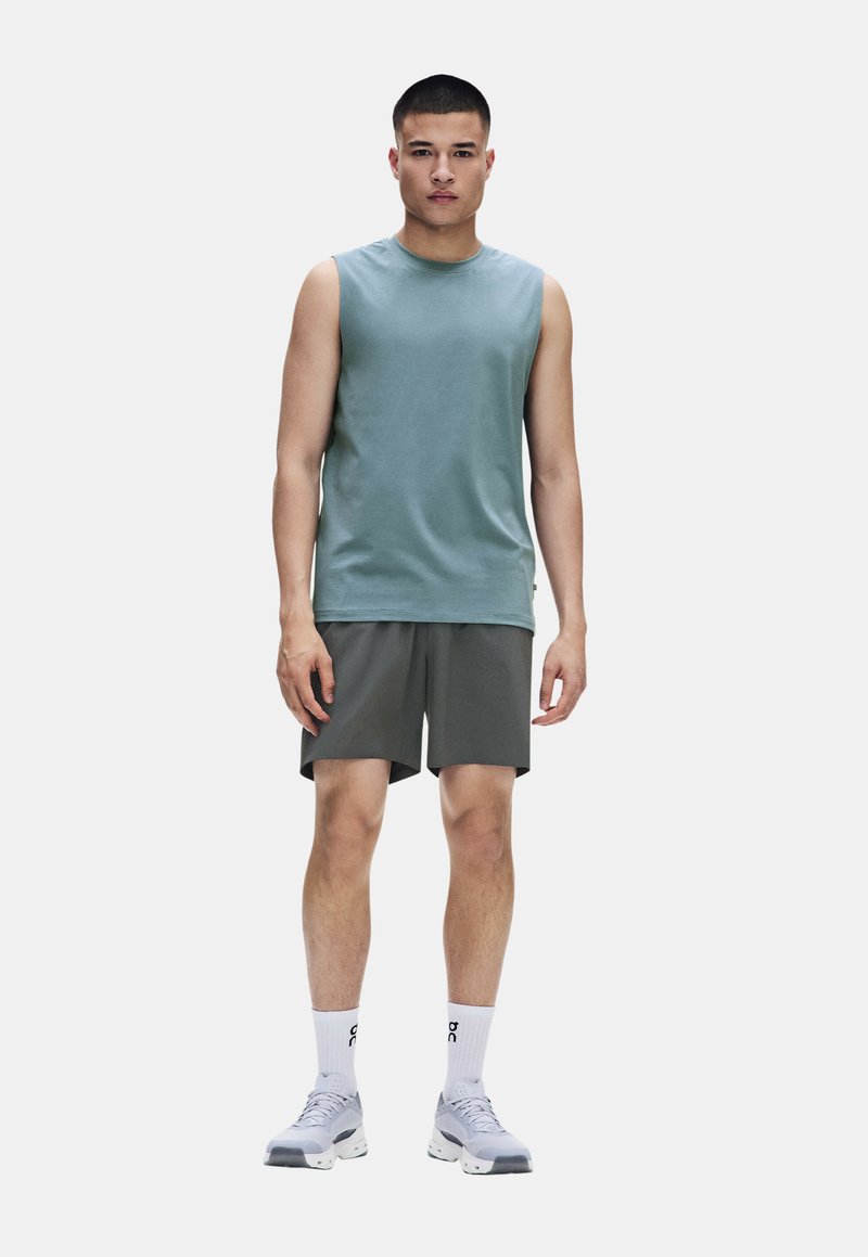 Jeune homme debout portant un débardeur turquoise, un short gris foncé, des chaussettes blanches montantes et des chaussures de sport blanches sur un fond uni.