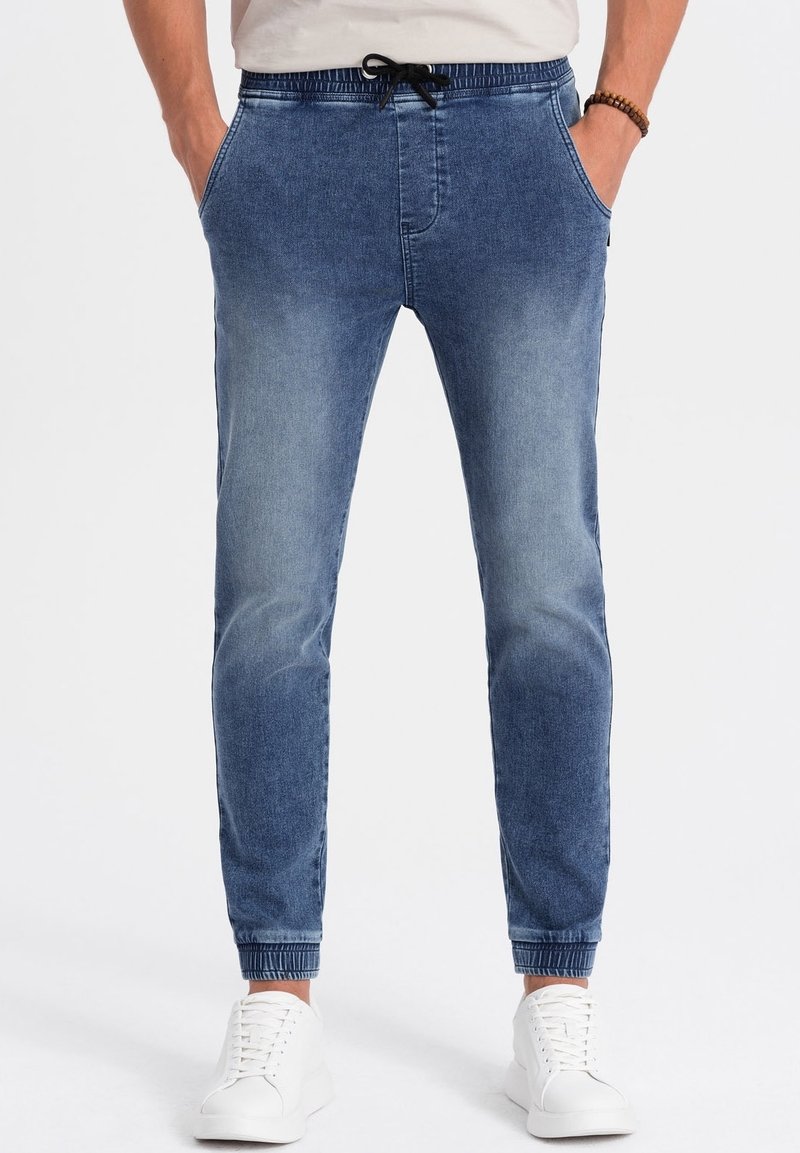 Homme portant un pantalon jogger en jean bleu avec des poignets élastiques et un cordon noir, les mains dans les poches, et des baskets blanches.