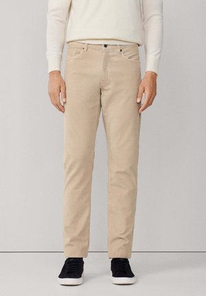 Mann trägt beige gerade geschnittene Hose, weißen Pullover und schwarze Sneakers, steht vor einfarbigem Hintergrund.
