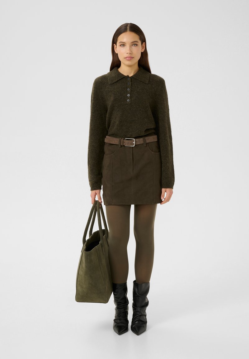 Olivgrünes Strickpoloshirt mit Kragen und Knopfdetails, kombiniert mit einem passenden Wildlederrock, grünen Strumpfhosen und schwarzen gerafften Stiefeletten.