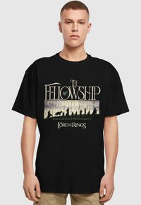 T-shirt en coton noir avec le texte "La Communaut�é" et "Le Seigneur des Anneaux", accompagné d'un graphique stylisé représentant des silhouettes dans un paysage.