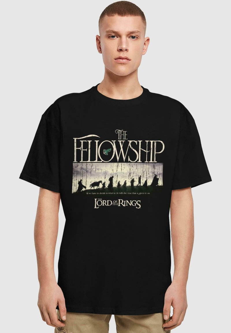 T-shirt en coton noir avec le texte "La Communauté" et "Le Seigneur des Anneaux", accompagné d'un graphique stylisé représentant des silhouettes dans un paysage.