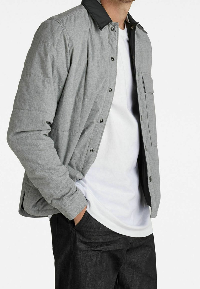 Grau gesteppte Jacke mit schwarzem Kragen und Knopfverschluss, ausgestattet mit einer Brusttasche und seitlichen Einschubtaschen, kombiniert mit einem weißen Hemd und dunklen Hosen.