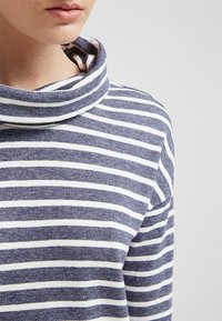 Långärmad skjorta med duskhals, tillverkad av texturerad marinblått tyg med vita horisontella ränder. Närbild av kragen och det övre tyget.