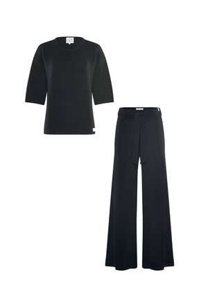 Young woman with red hair stands barefoot against a plain background, wearing a black ribbed top with three-quarter sleeves and wide-leg pants.