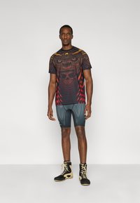 Μαύρο γραφικό μπλουζάκι με κόκκινα και πορτοκαλί σχέδια· συνδυασμένο με εφαρμοστά σκούρα σορτς και ψηλά μαύρα και μπεζ παπούτσια.