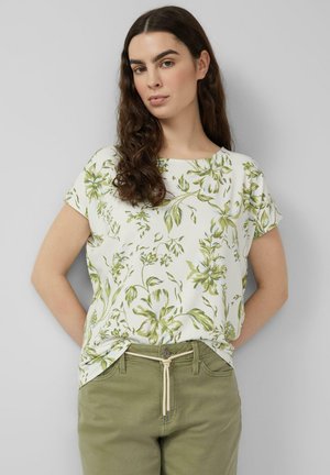 Kobieta o długich ciemnych włosach, ubrana w białą bluzkę z krótkim rękawem z zielonym kwiatowym wzorem oraz oliwkowe spodnie z paskiem z troczkami.