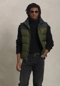 Olivgrön pufferväst med mörkgrå axlar, med en dragkedjaficka, bärs över en svart kabelstickad tröja och svarta jeans. Kompletteras med solglasögon.