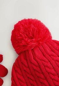 Röd stickad mössa med en stor pom-pom på toppen, prydd med ett texturerat kabelmönster. Mjuk garn med en chunky design. Mindre röda handskar i närheten.
