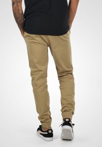 Kaki tapered bukser med elastiske mansjetter, laget av bomullsblanding. Brukt med svarte og beige sneakers, som viser et rent, uformelt design.