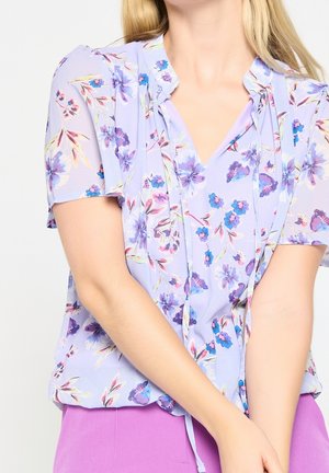 Femme portant un chemisier violet clair avec des motifs floraux bleus et roses et des manches courtes, associé à un pantalon violet vif.