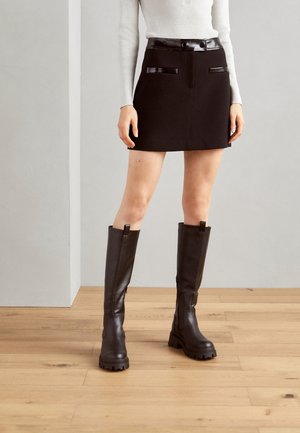 Femme portant une mini-jupe noire avec un bord brillant et de grandes bottes noires épaisses, debout sur un sol en bois contre un mur gris.