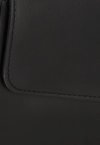 Sort lærertaske med glat tekstur, kontrastsyetninger og flaplukning. En subtil glans fremhæver dens elegante design og kanter.