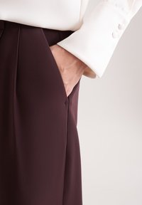 Bordeauxkleurige broek met een zachte textuur, voorzien van een zijzak, gecombineerd met een witte blouse met knoopmanchetten.