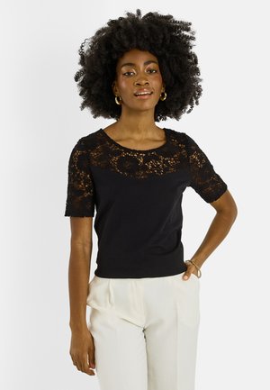 Mujer con pelo rizado natural que lleva pendientes de aro dorados, parte superior negra de encaje de manga corta, pantalones blancos y una pulsera dorada, de pie con la mano en la cadera.