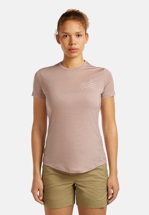 Junge Frau mit lockigem Haar im Pferdeschwanz, die ein mauvefarbenes Kurzarmshirt mit weißem Berglogo und olivgrüne Shorts trägt.