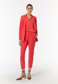 Damski garnitur skrojony w jasnym koralowym kolorze, składający się z dopasowanej marynarki i zwężanych spodni, zestawiony z bluzką z dekoltem w serek. Brązowe buty na obcasie.