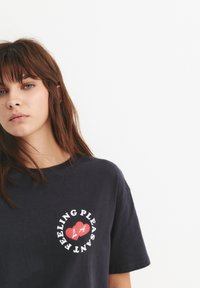 Tricou din bumbac bleumarin, cu un grafic rotund ce conține textul "SENTIMENT PLĂCUT" și o inimă roșie. Mâneci scurte, croială lejeră.