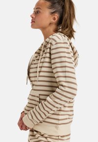 Sudadera a rayas beige con un bolsillo tipo canguro, cordones y puños acanalados. El material es suave, con un corte relajado y de grosor medio.