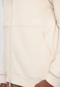Veste beige zippée avec une poche et le logo blanc EA7 Emporio Armani sur la poitrine, portée par une personne à la peau brune.