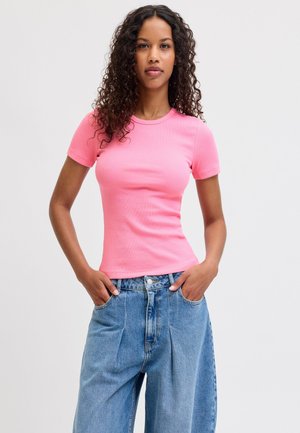 Jeune femme aux cheveux bouclés portant un t-shirt moulant rose à manches courtes et un jean bleu ample, les mains dans les poches, sur un fond uni.