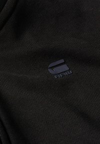Tela de algodón negra con una textura acanalada que presenta un logotipo en azul oscuro y el texto "RAW". No hay patrones ni detalles adicionales.