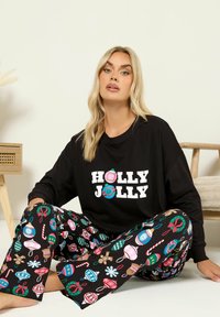 Musta pitkähihais toppi, jossa on teksti "HOLLY JOLLY"; värikäs juhlava kuosi löysissä mustissa pyjamahousuissa, joissa on koristeita ja karkkikeppejä.