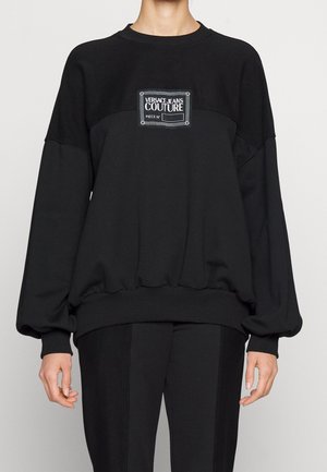 Sweat-shirt noir oversize à manches longues avec un patch logo Versace Jeans Couture sur la poitrine, porté avec un pantalon noir.