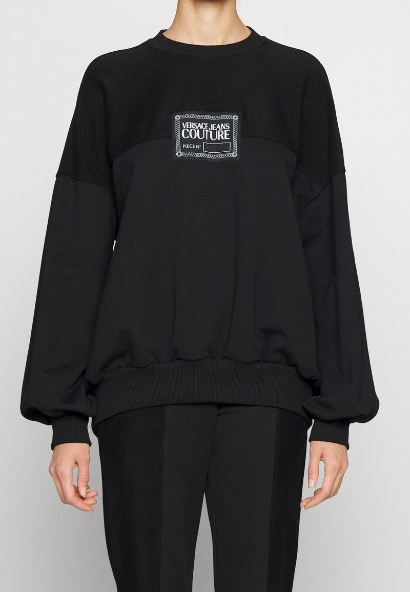 Sweat-shirt noir oversize à manches longues avec un patch logo Versace Jeans Couture sur la poitrine, porté avec un pantalon noir.