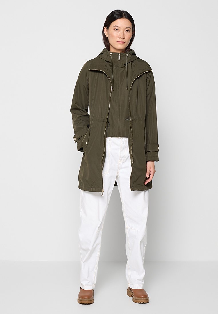 Kobieta w oliwkowozielonym bejsbolówce z kapturem, białych spodniach z wysokim stanem i brązowych botkach na kostkę, stojąca na tle jasnoszarym.