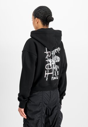 Frau mit geflochtenem Dutt, die einen schwarzen Hoodie mit weißem Blumen- und Textdesign trägt, kombiniert mit schwarzen gerafften Hosen, steht vor weißem Hintergrund.