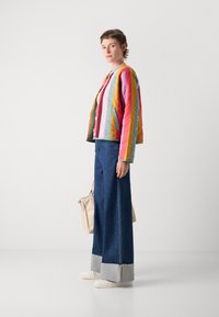 Bunte wattierte Jacke mit vertikalen Streifen in Pink, Gelb und Blau; kombiniert mit weit geschnittenen Jeanshosen und weißen Sneakers.